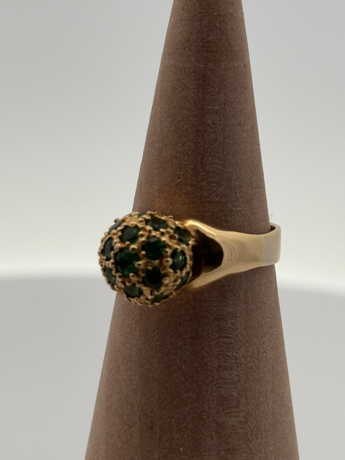 Gold ring with green gemstones on a brown stand against a white background