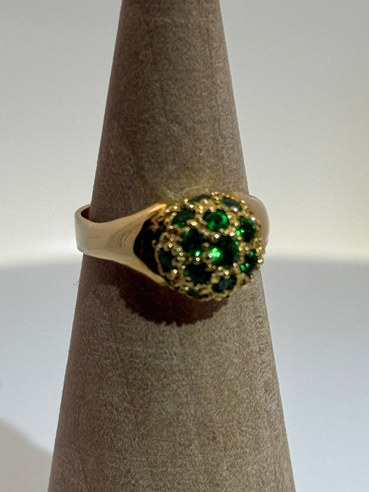 Gold ring with green gemstones on a brown cylindrical stand against a white background