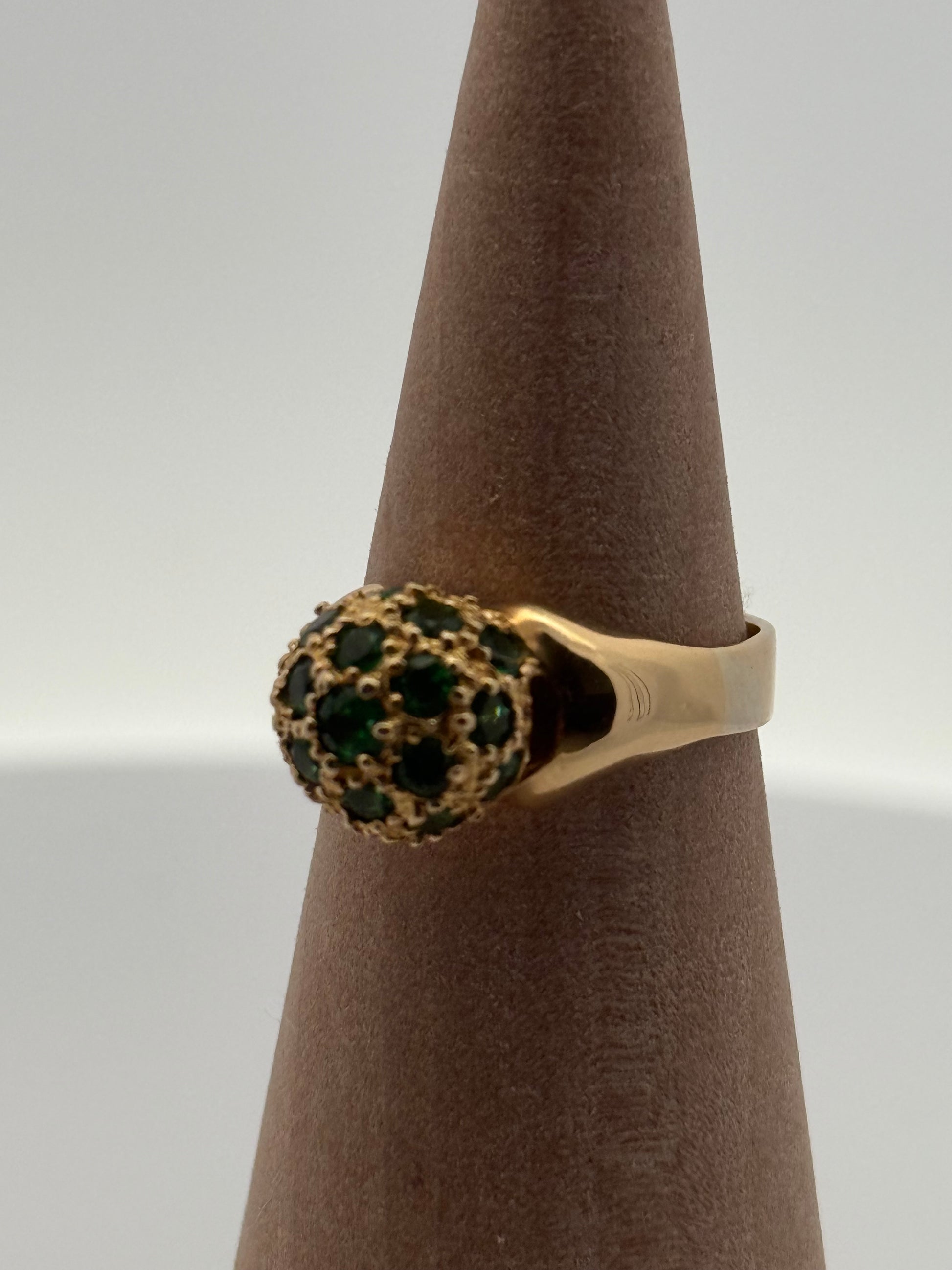 Gold ring with green gemstones on a brown stand against a white background